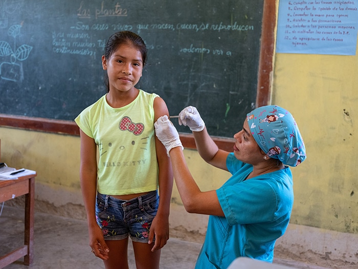 A health work vaccinates a girl in Peru in front of a chalkboard.