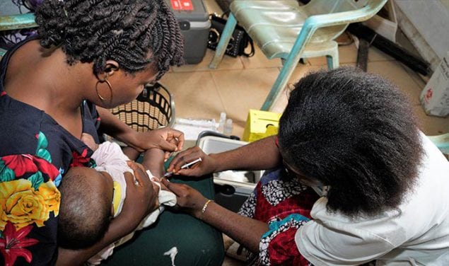Woman holds infant receiving a vaccine by a healthcare worker in Nigeria, 2021.