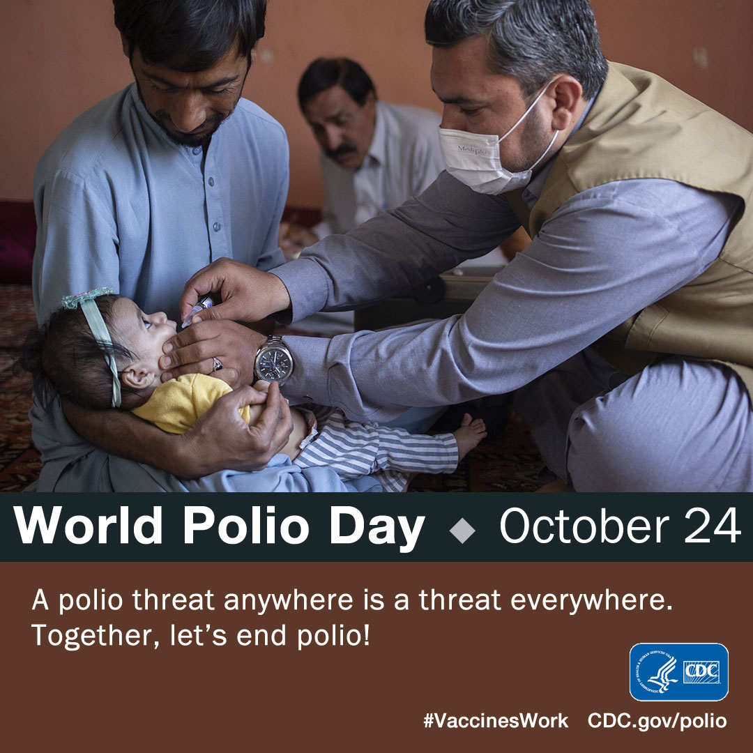 A man holds an infant while sitting an a carpet while another man squeezes drops of oral polio vaccine into the baby's mouth.