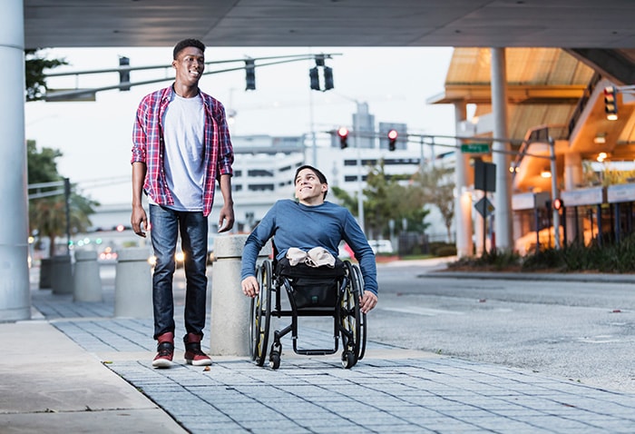 Two men talking while walking down the street. One man is in a wheelchair.