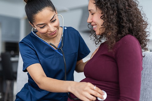 Physician wearing navy blue scrubs holding a stethoscope to a pregnant person’s belly.