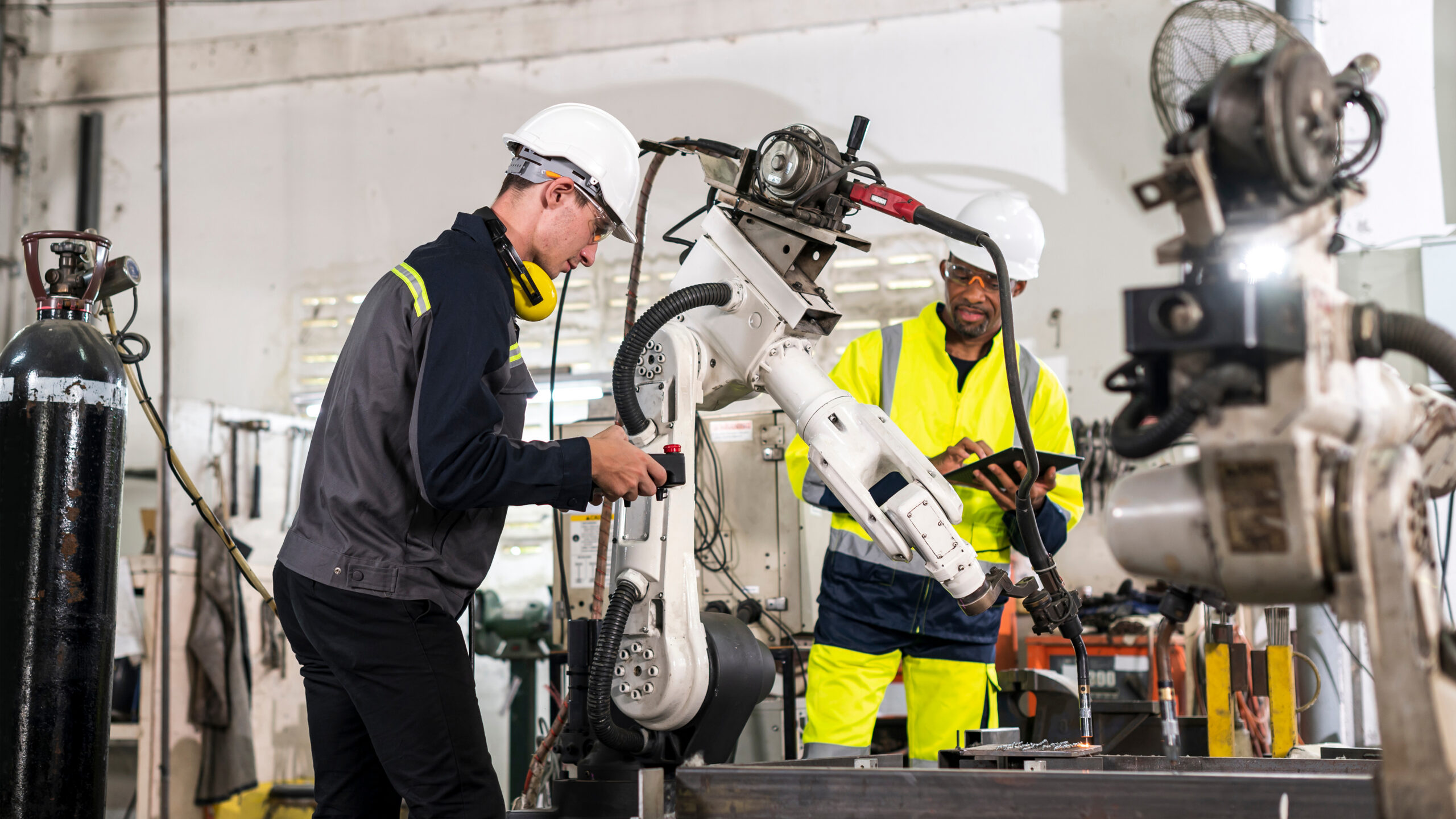 Two industrial workers in hard hats and safety gear operate a large robotic arm in a manufacturing workshop, with machinery, tools, and equipment visible in the background.