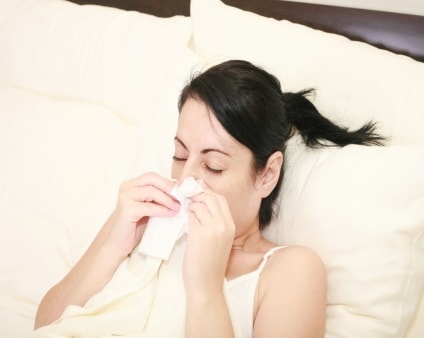 Woman at home sick in bed covering her cough with a tissue.)