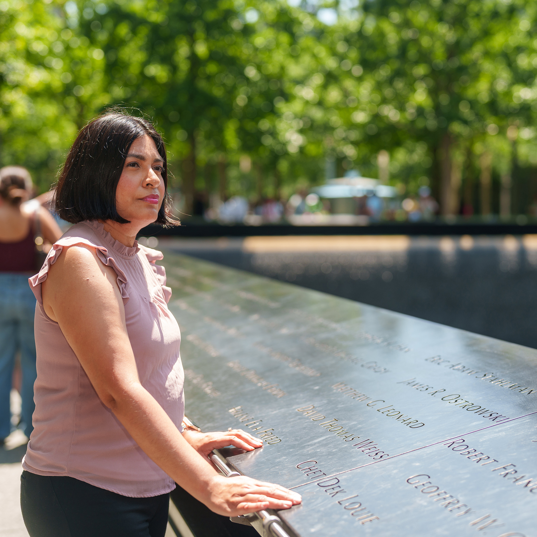Woman standing at the 9/11 Memorial plaza, resting her hand on engraved names.