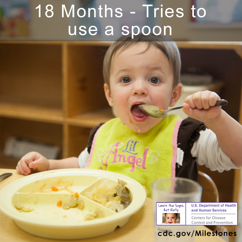 The boy in this photo is trying to use a spoon while wearing a yellow bib.
