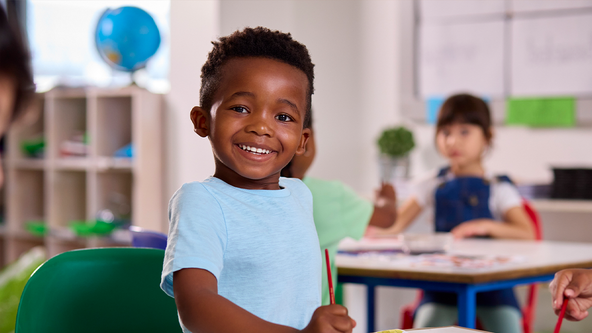 4 year old smiles at a desk in a classroom.
