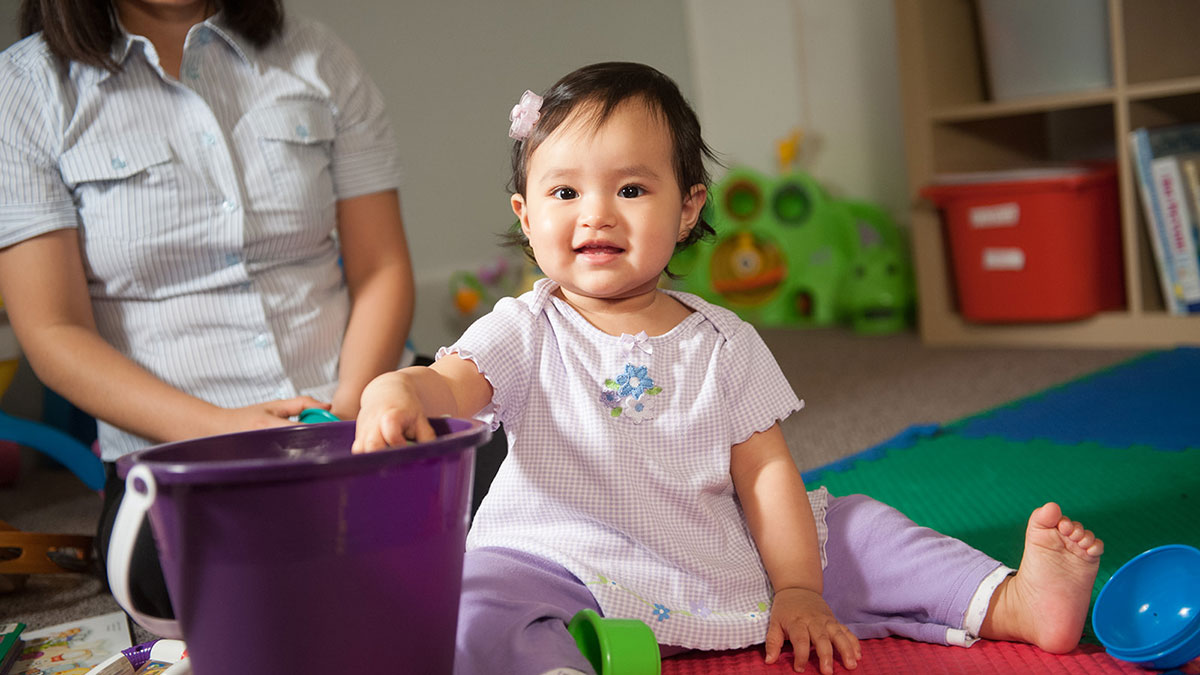 A baby girl sits on the floor of a room with toys and a bucket, a child care professional watches nearby.