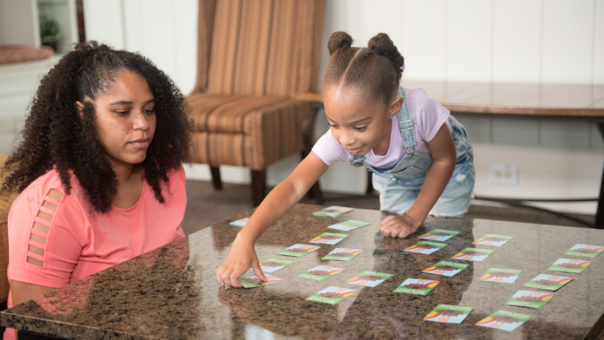 5 year old girl turns over a card on the table, her mom sits next to her.