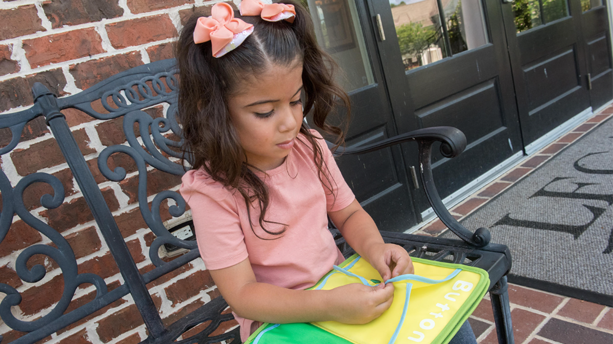 Little girls sits on a bench and buttons two pieces of fabric together in a book.