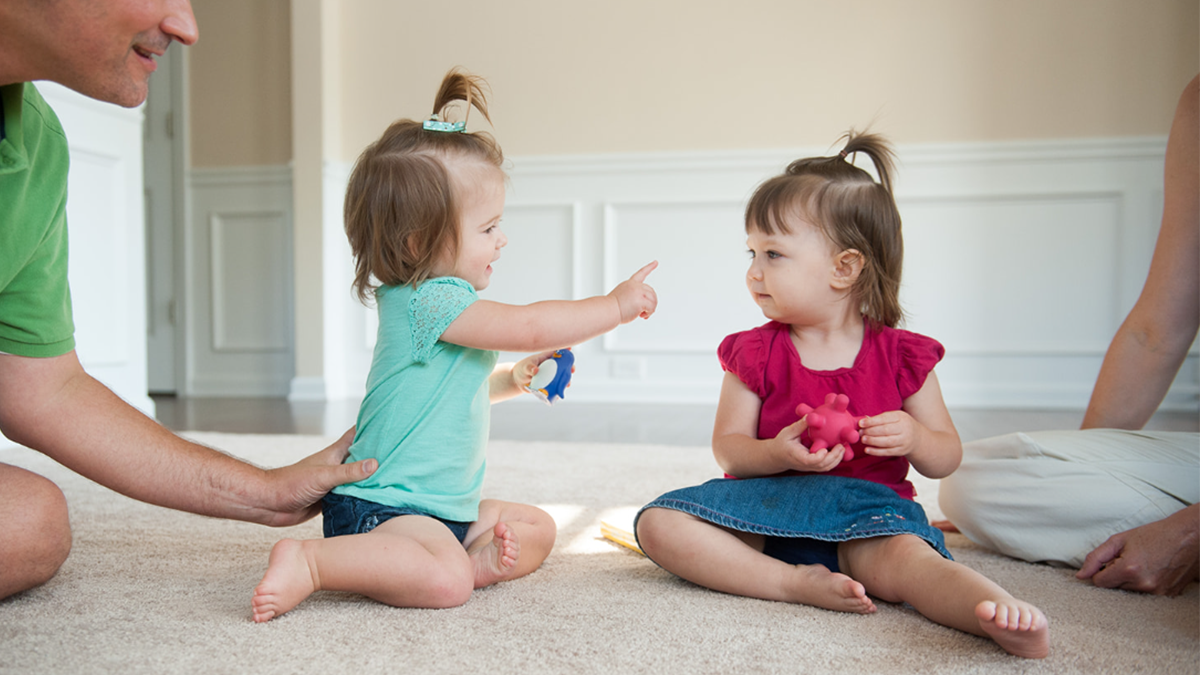 Two baby girls sit on the floor playing with toys in their hands