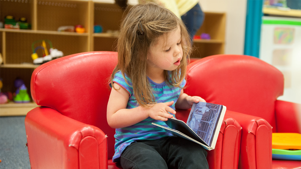 Little girl sits in a chair and turns pages in a picture book.