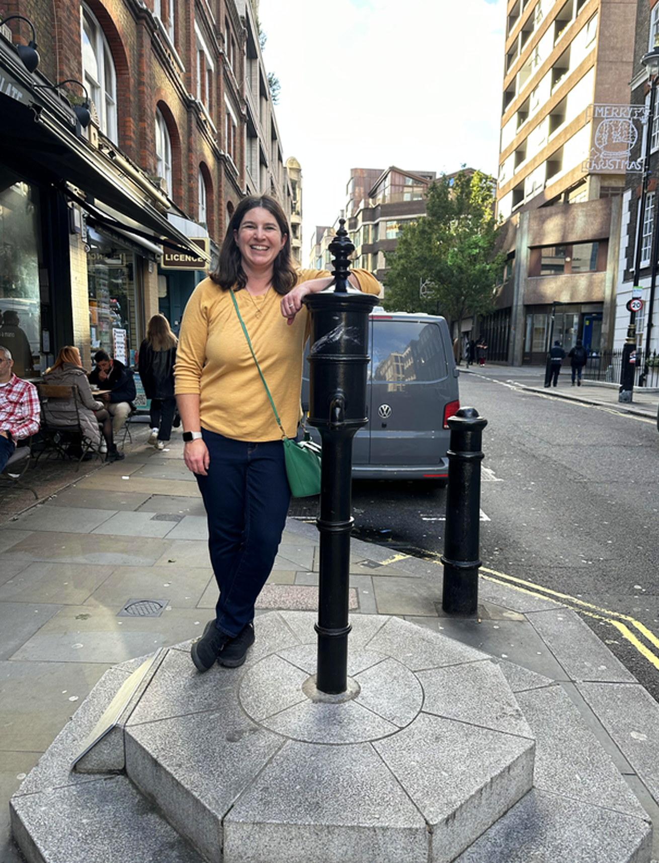 Smiling woman leaning against a vintage water pump embedded into a octagonal platform in the sidewalk of a London street