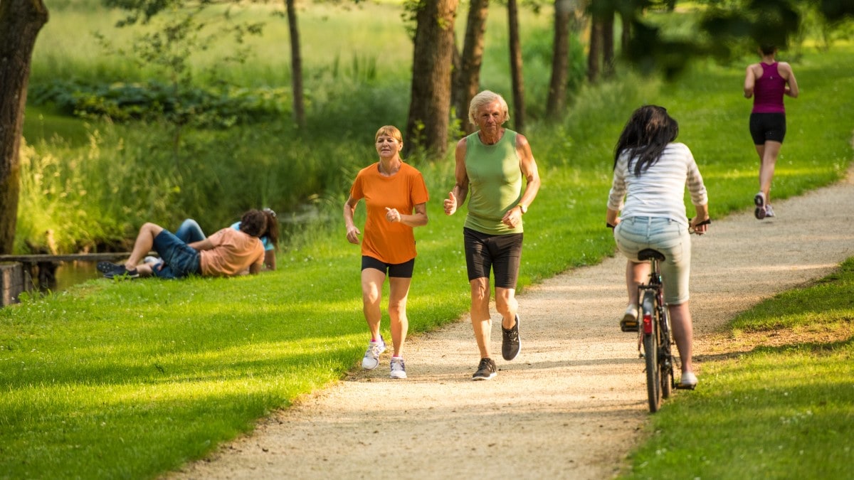 Older couple running in a park, with people laying by a lake and a lady biking on a path.