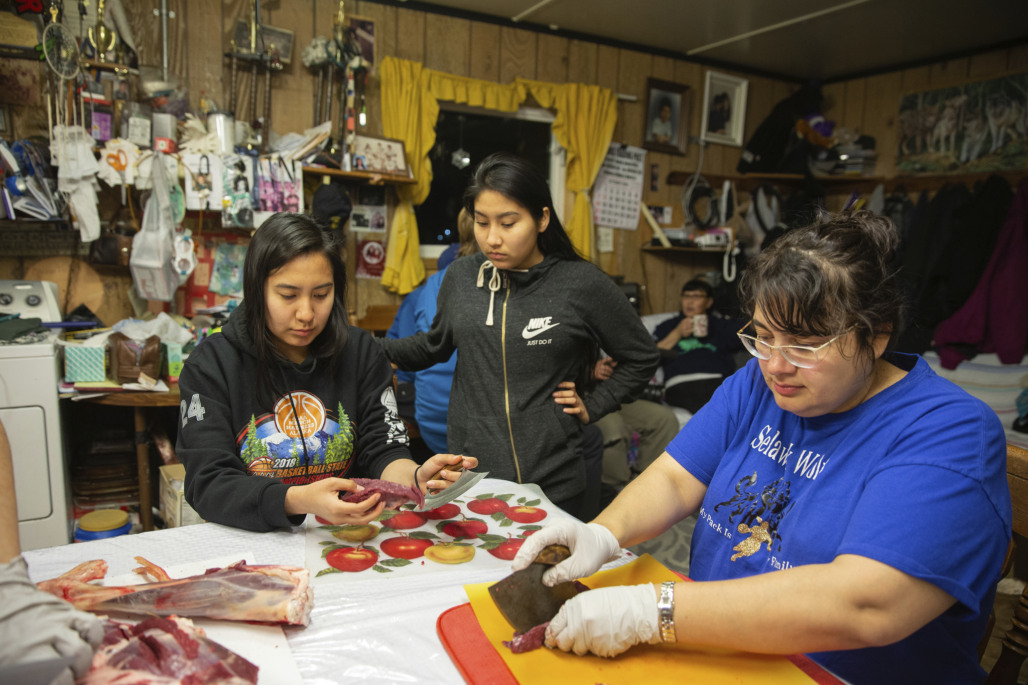 Two women seated at a table chopping food, while a younger woman stands at the table.