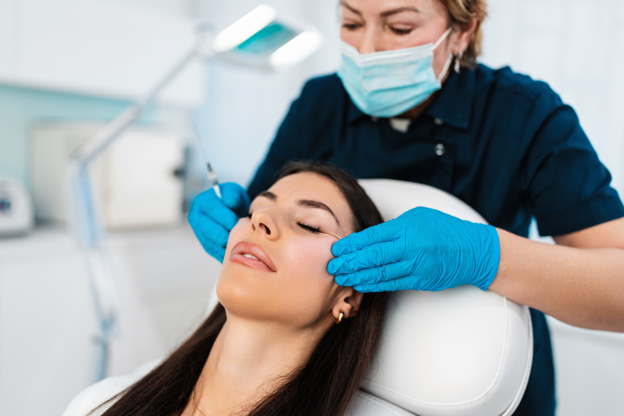 Provider wearing gloves and mask prepares to give cosmetic injections to a patient, who is leaning back in a chair.