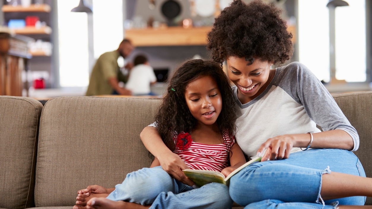 A mom and daughter sit on a sofa and read a book together.