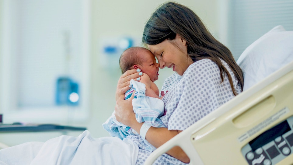 A new mom sits in a hospital bed and holds her newborn in front of her. She is wearing a hospital gown.