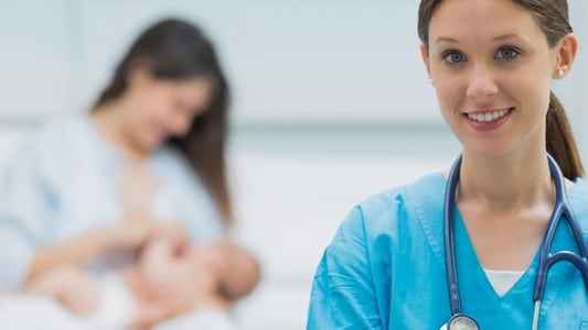 Mother breastfeeding an infant in the background with a healthcare professional in foreground.