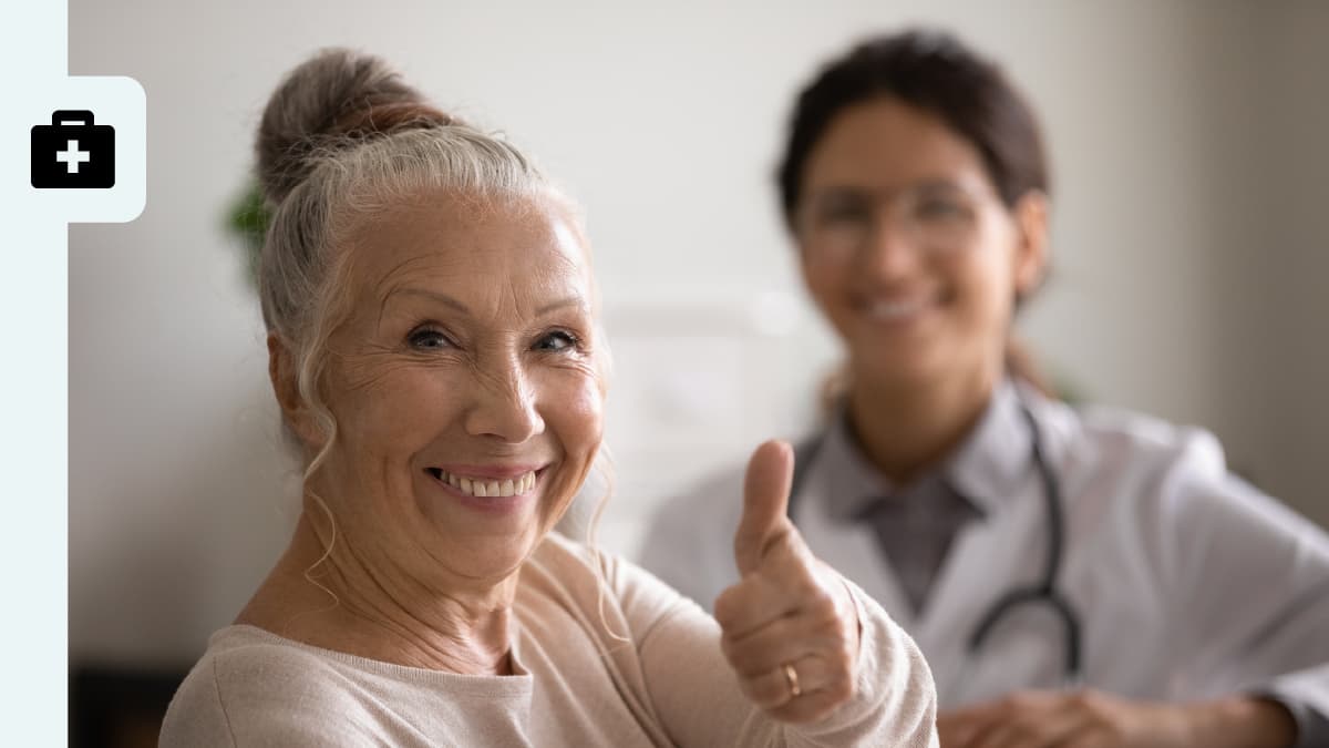 An older woman smiles and gives a thumbs up sign as her doctor looks on