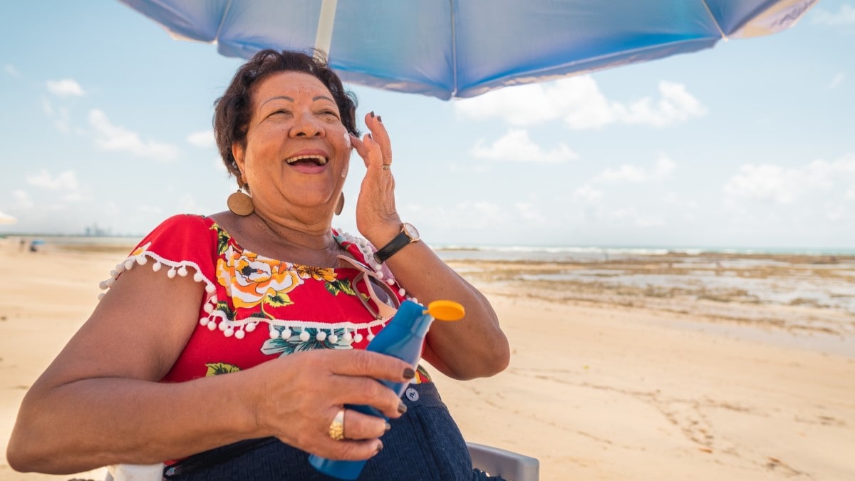 Foto de una mujer mayor sentada debajo de un paraguas en la playa, aplicándose filtro solar