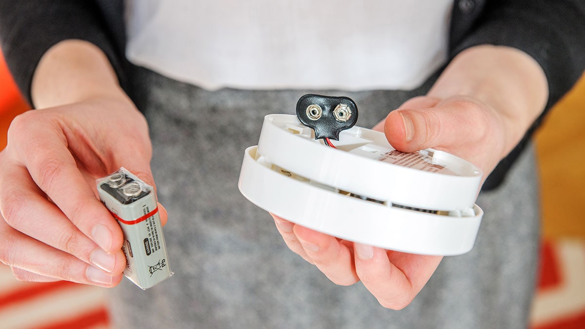 A person holding a carbon monoxide alarm in one hand and a battery in the other hand