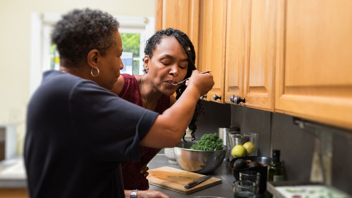 Slightly older woman giving a spoonful of what's cooking to younger woman.