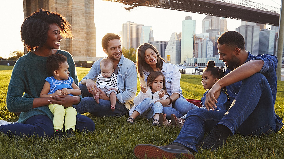 Several diverse, young parents with their children, sitting in a park with the cityscape in the background.