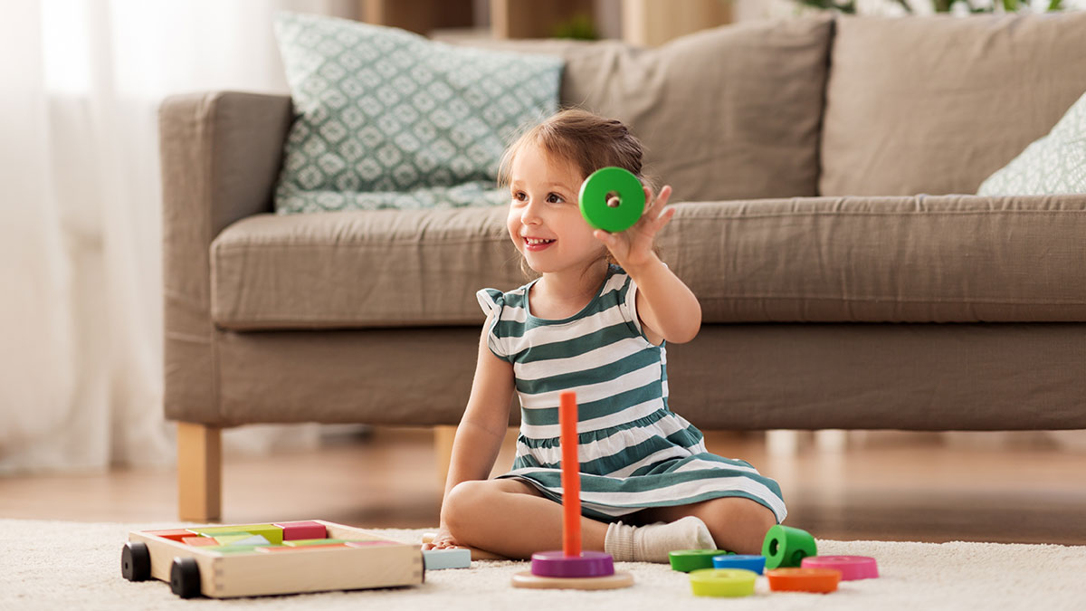 Smiling toddler sitting on the floor, holding a green ring while playing with a colorful stacking toy.