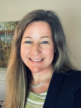 A headshot of Dr. Lias. She has long blonde hair and is wearing a black suit jacket with a green and white striped shirt.