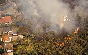 A top-down view of a fire in a wooded area, creating a large amount of smoke near buildings and residencies.