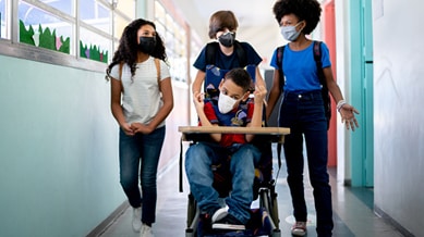 A group of teenagers walk down a school hallway, and one student is being pushed down the hall in his wheelchair. Everyone is wearing facemasks.