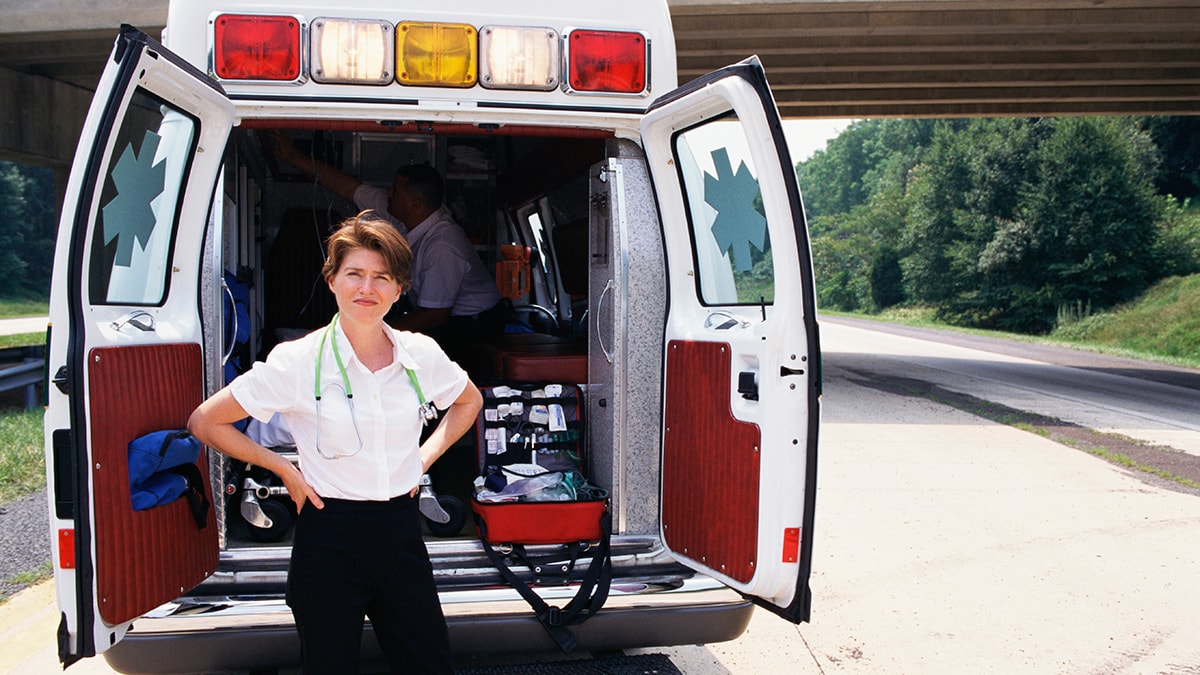 An Emergency Medical Services technician standing outside the back of an ambulance with its doors open.