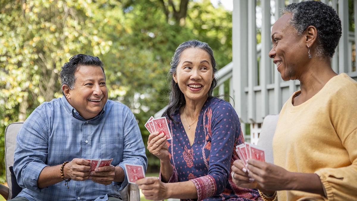 Three older friends, one man and two women, playing cards together and smiling.