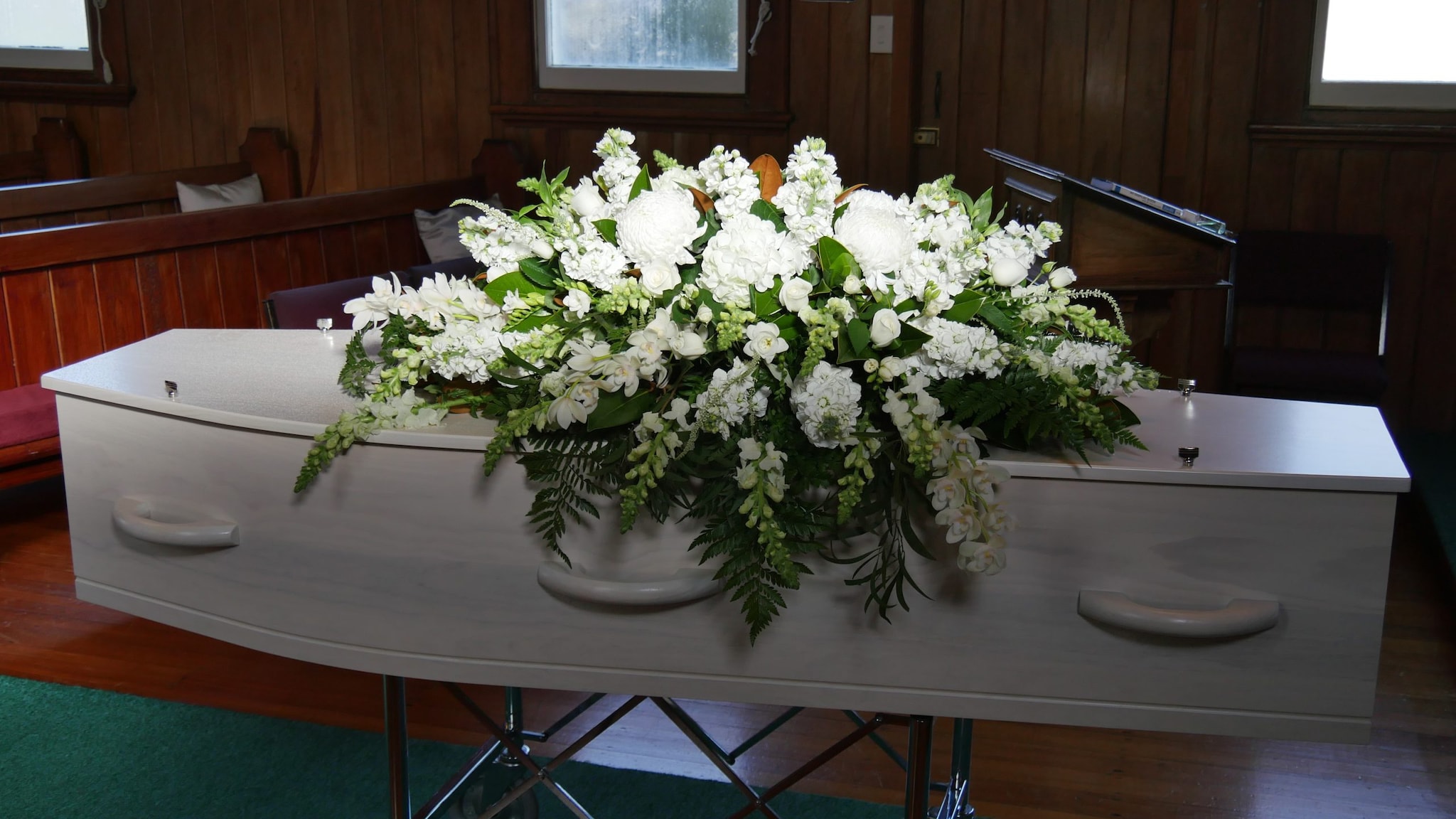 Image shows a white casket sitting on a raised holder. It has a large spray of white flowers atop it and light shining down on it.