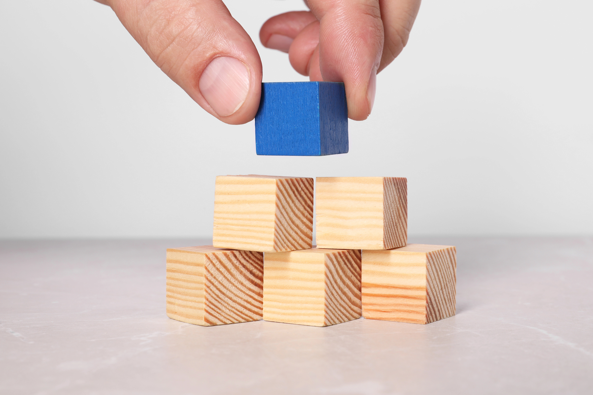 A hand puts small blue block onto stack of five wooden blocks