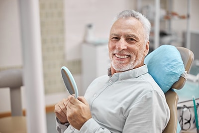 Hombre mayor sonriente sentado en el sillón del dentista con un espejo en la mano