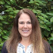 Headshot of Angela Weaver outdoors in front of a leafy tree. She has brown hair and a white top.