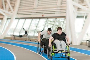 Two people racing in wheelchairs on an indoor track. The person in a black shirt is ahead of the person in a grey shirt.