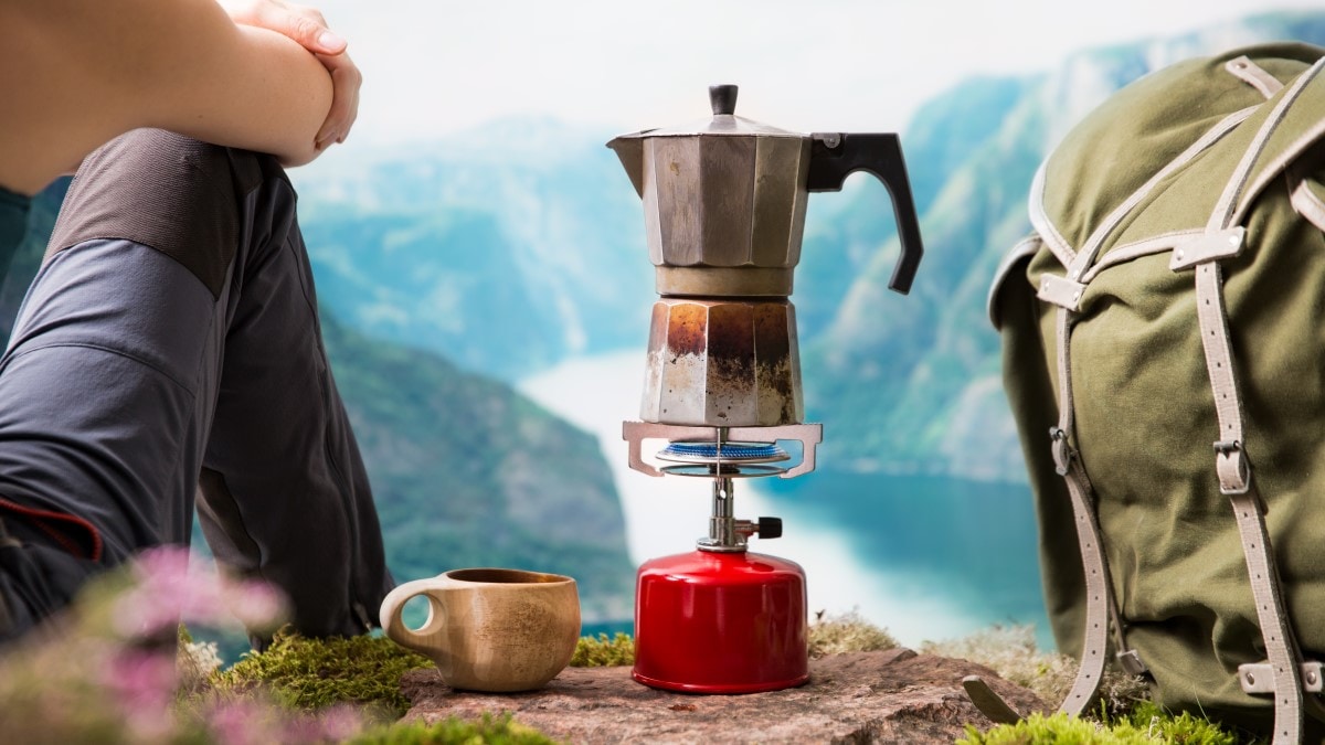 Hiker sitting on a rock looking out over mountains and a water body. A mug, small kettle boiling over a gas burner, and backpack are next to the hiker on the rock.