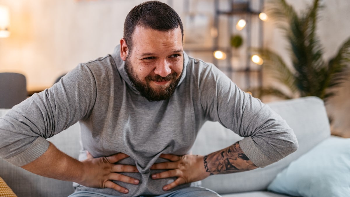 Man with a pained expression sitting on a couch clutching his stomach with both hands