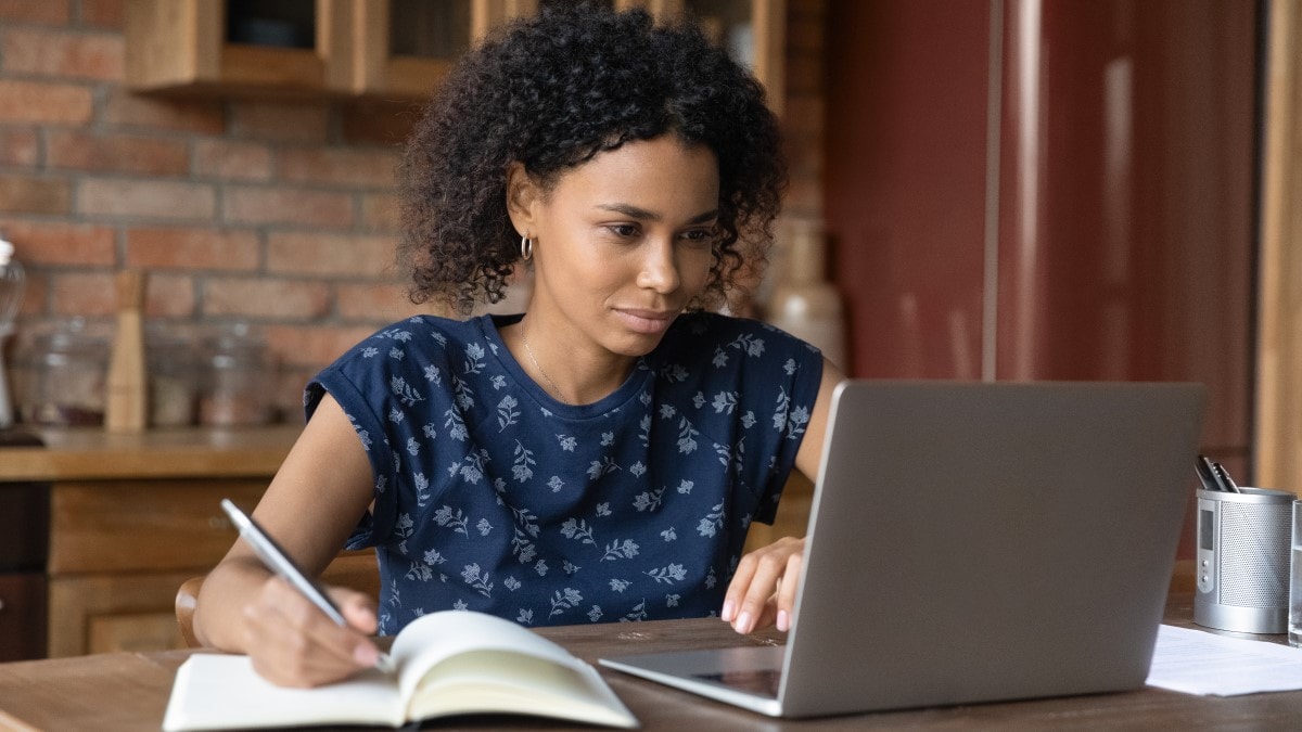 Woman in her kitchen looking at a laptop and writing in a notebook