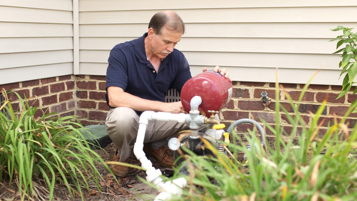 Person squatting down to check a well pump outside a house