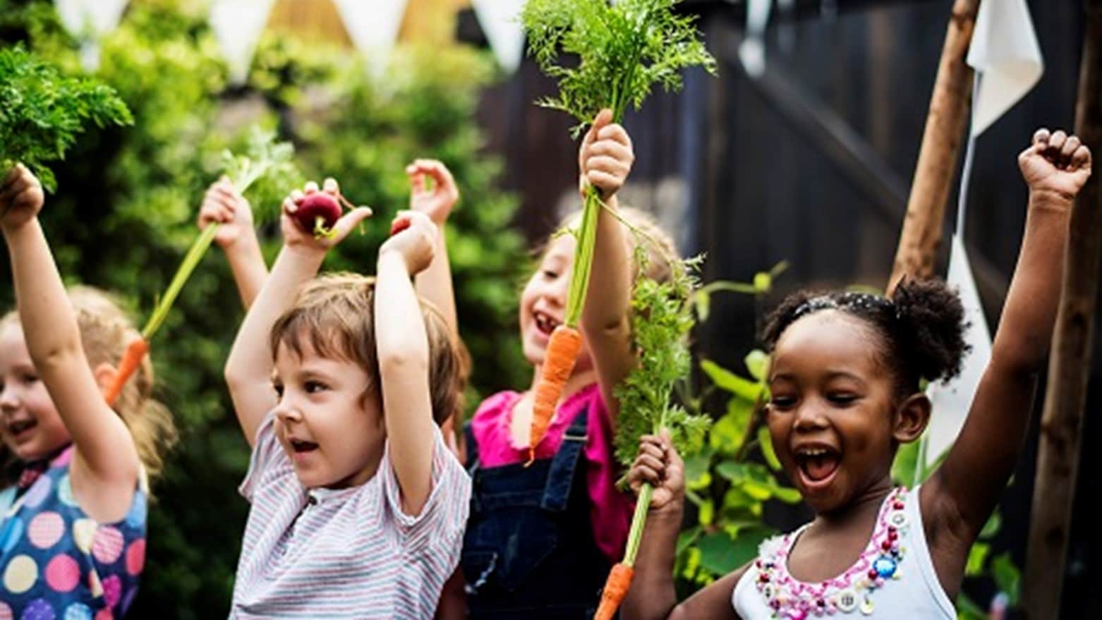 Young children in a garden and holding carrots over their heads.