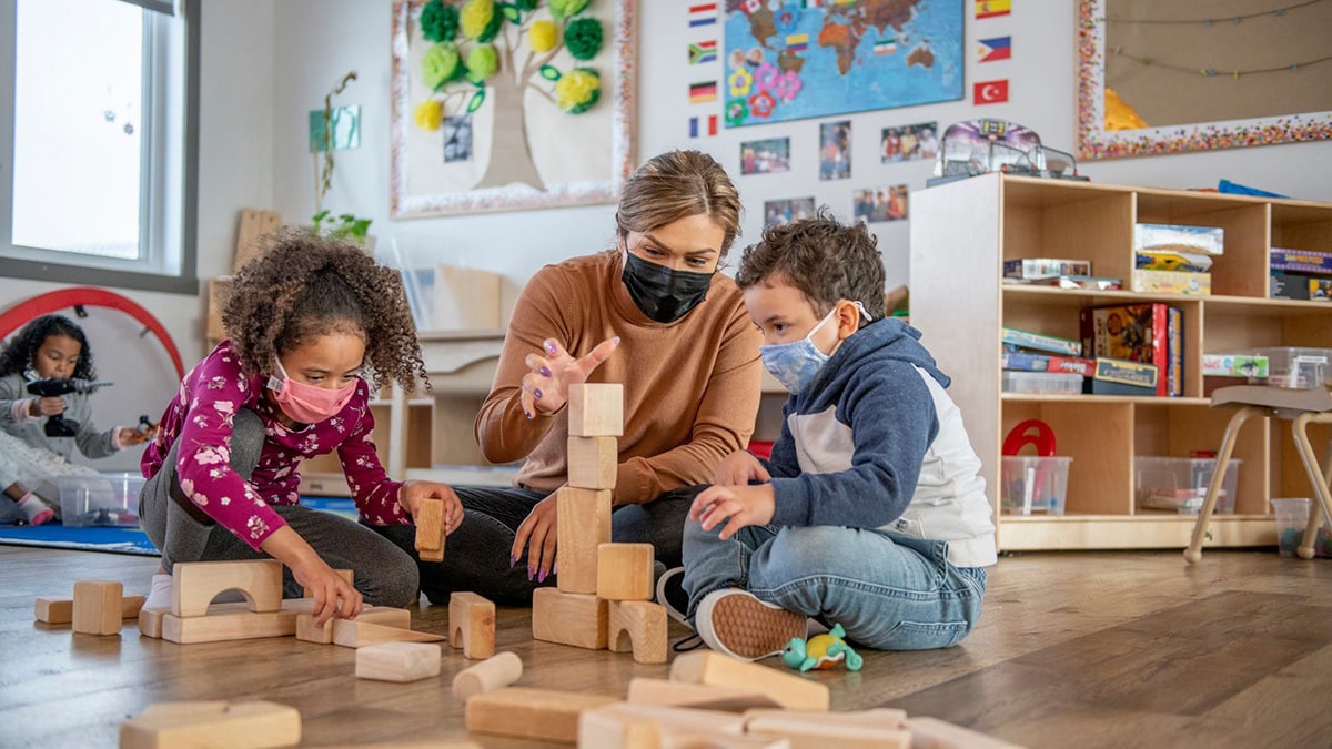 An early educator wearing a protective face mask while playing building blocks with children.