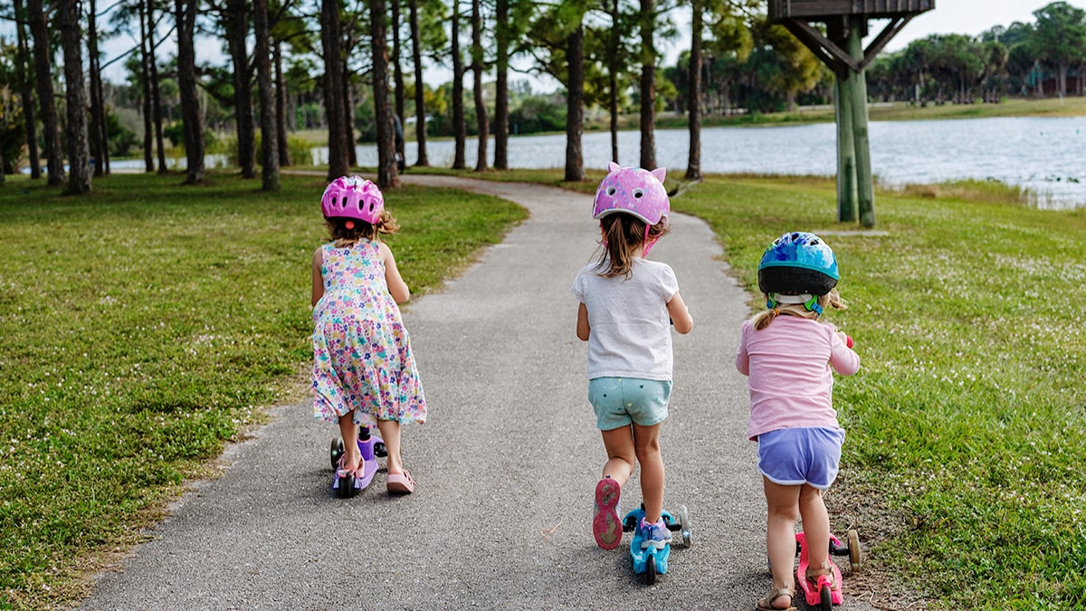 Three young children using scooters in a park wearing safety helmets