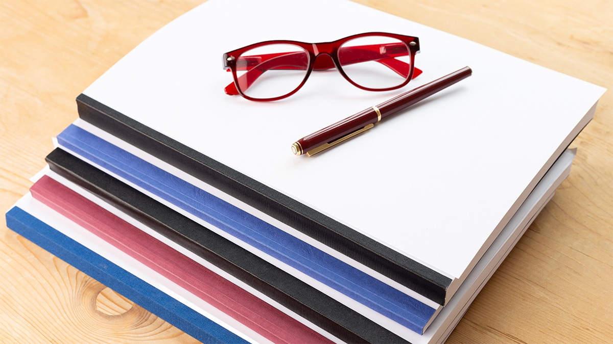 Stack of publications on a desk with a pair of glasses and a pen.