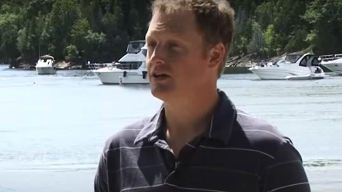 Man in blue shirt speaking wih lake and boats in background.