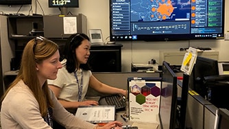 Two women sitting at a desk with a data screen in the background.