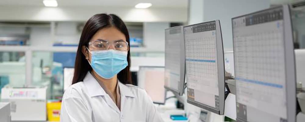 Masked laboratory worker in lab looking straight ahead with computers that feature data to the side.