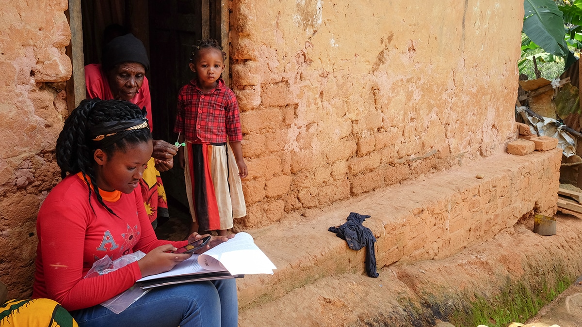 Community health worker sitting outside home, writing in notebook with woman and child looking on.